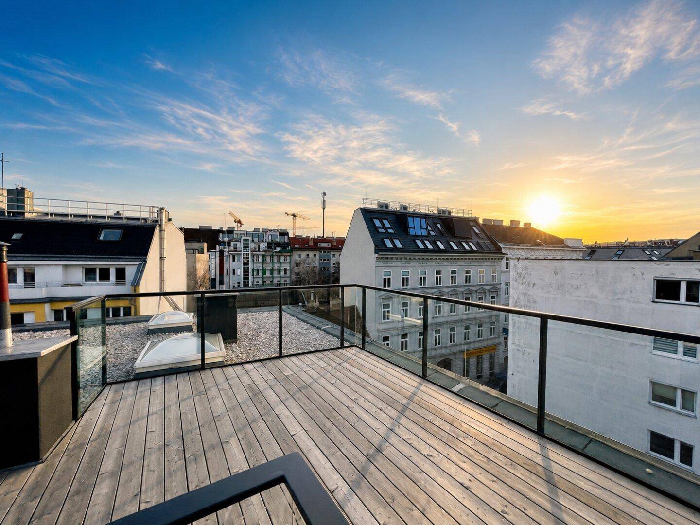 Dachterrasse mit Holzboden und Glasgeländer bei Sonnenuntergang, bietet weite Stadtblicke.