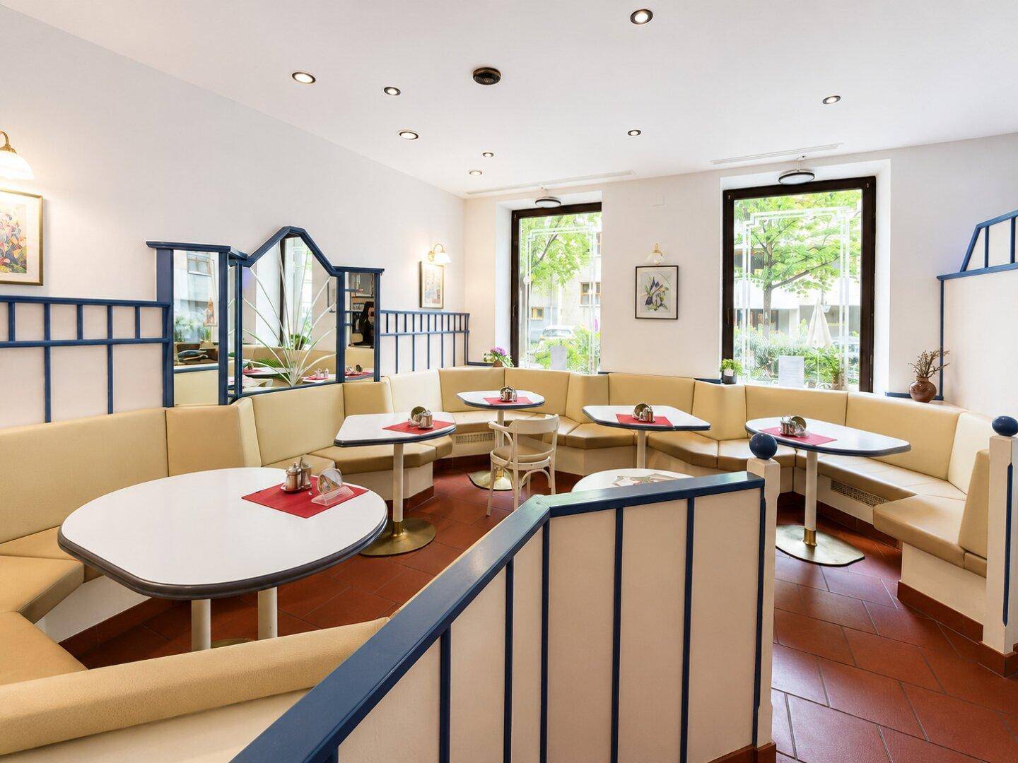 Heller Café-Innenraum mit beigen Sitzbänken, kleinen Tischen und blauen Akzenten, durch große Fenster gut beleuchtet.