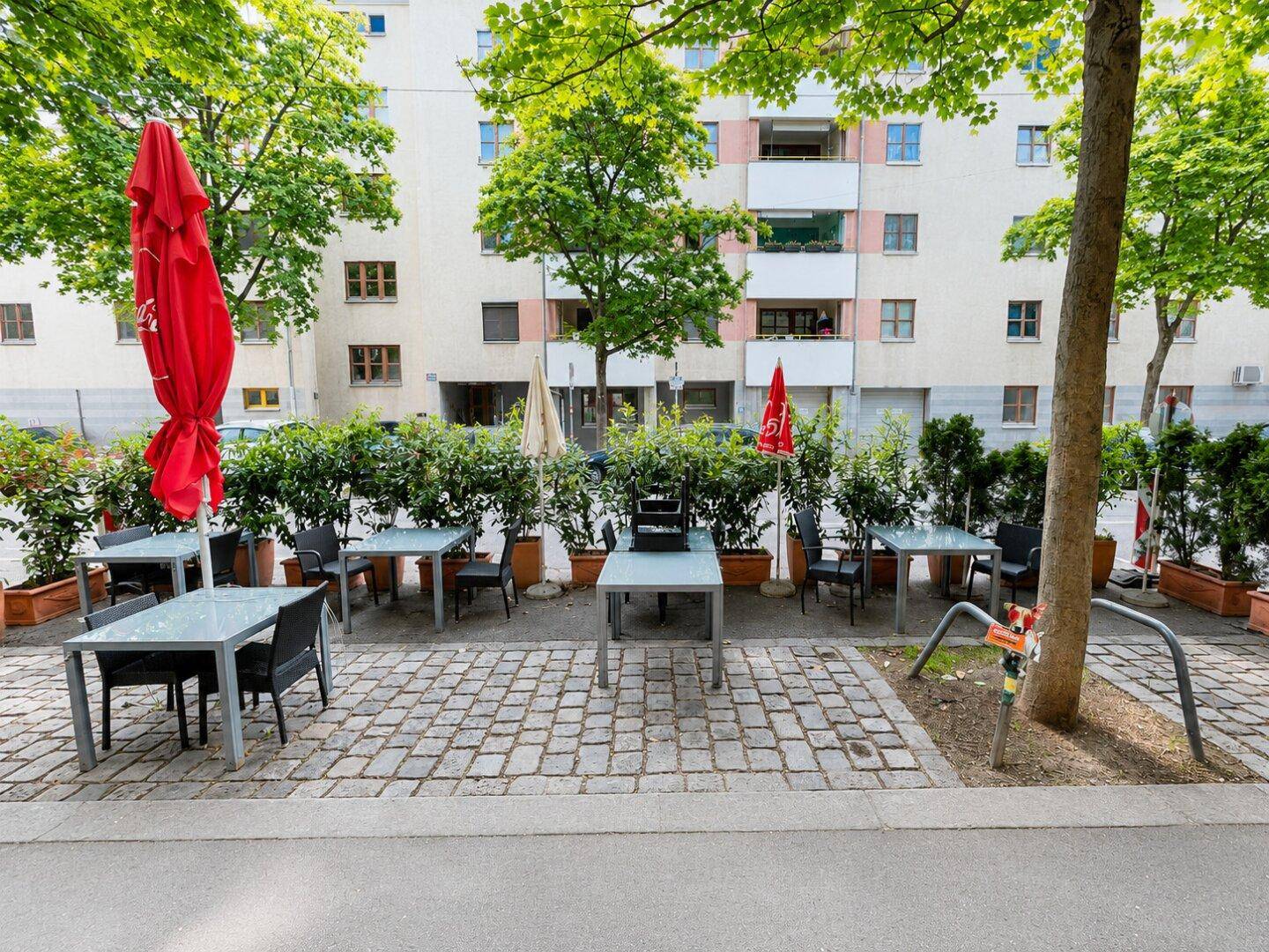 Außenbereich mit Tischen, Stühlen und roten Sonnenschirmen, durch Hecken von der Straße getrennt, vor einem Wohngebäude.