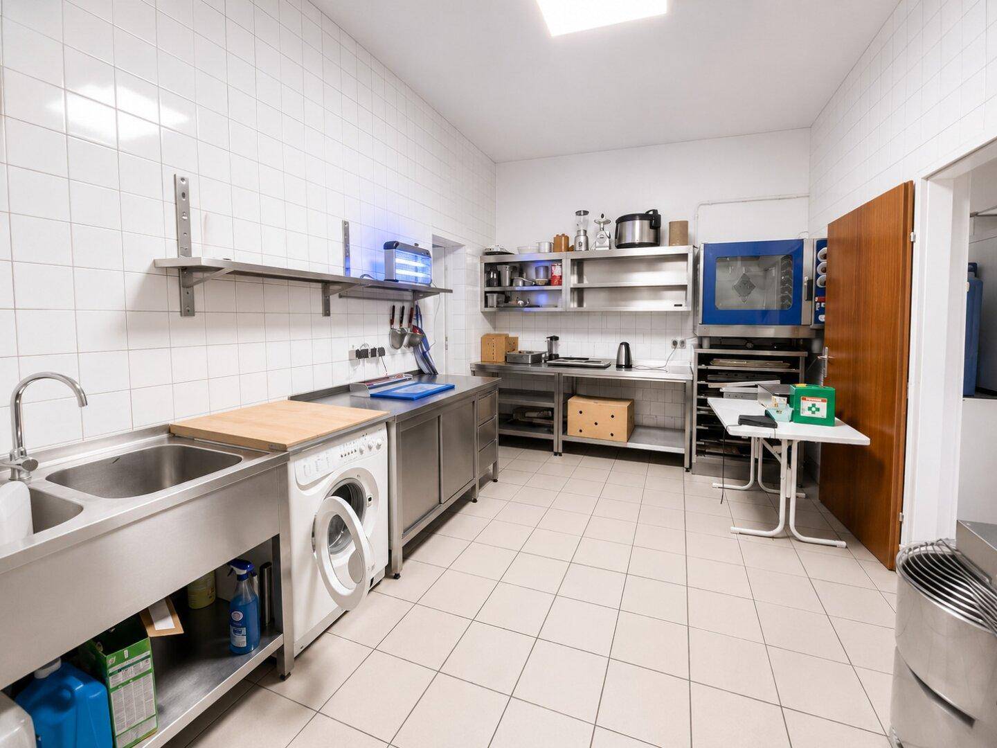Geräumige Gewerbeküche mit großem Spülbecken, Waschmaschine, Edelstahl-Arbeitsflächen und Fliesenboden.