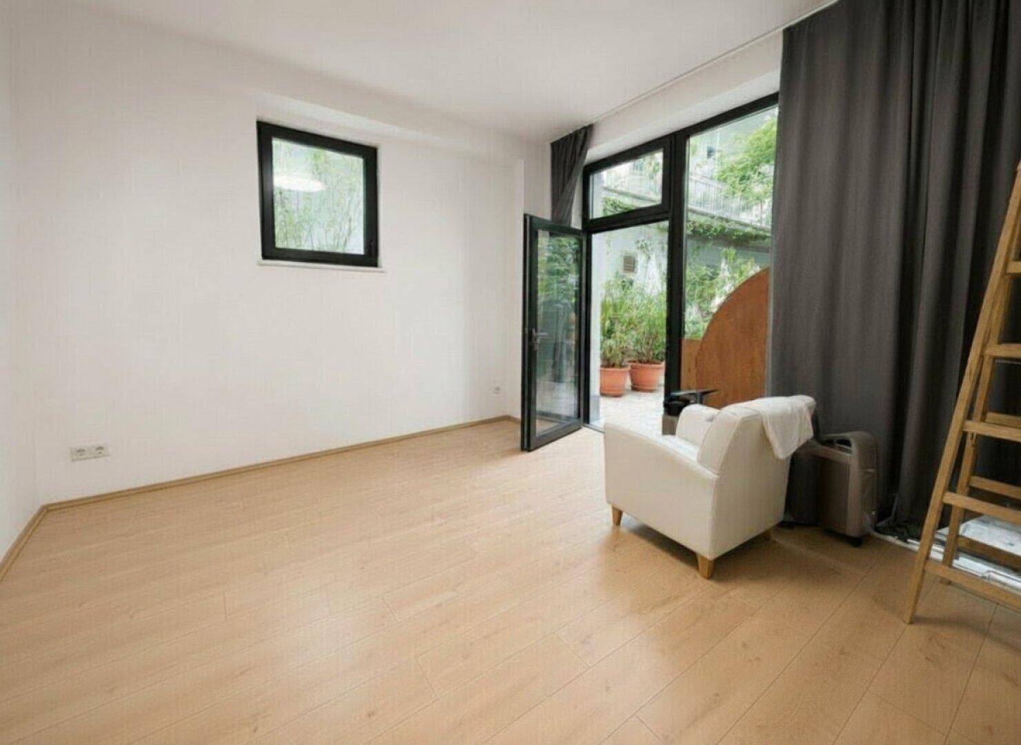 Heller, leerer Raum mit Laminatboden und großen Fenstern, die Zugang zu einem Außenbereich bieten.