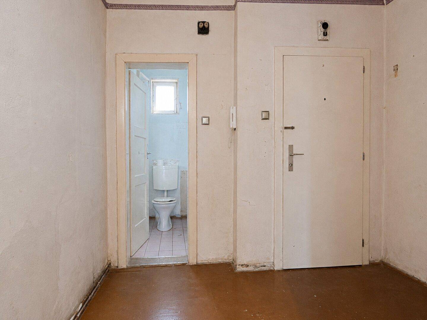 Kleiner Eingangsbereich mit Blick auf ein separates WC, alter Bodenbelag und einfache Wände.