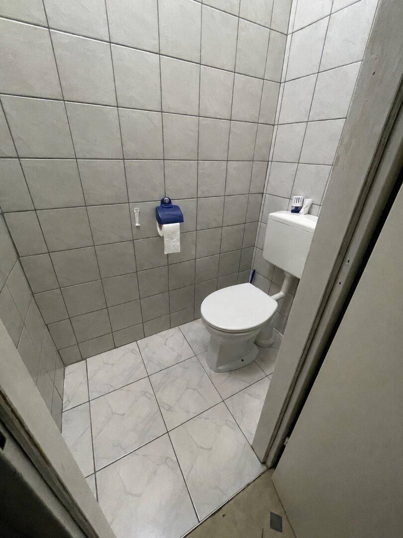 Einfache Toilette mit hellgrauen Fliesen an Wänden und Boden, funktional und sauber.