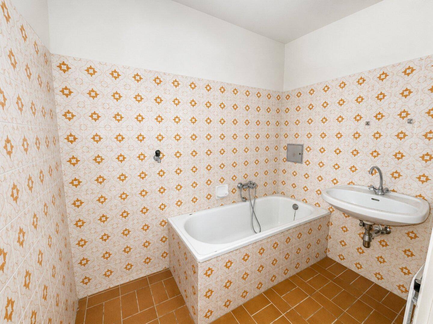 Badezimmer im Originalzustand mit Badewanne, Waschbecken und auffälligen orange-weißen Fliesen.