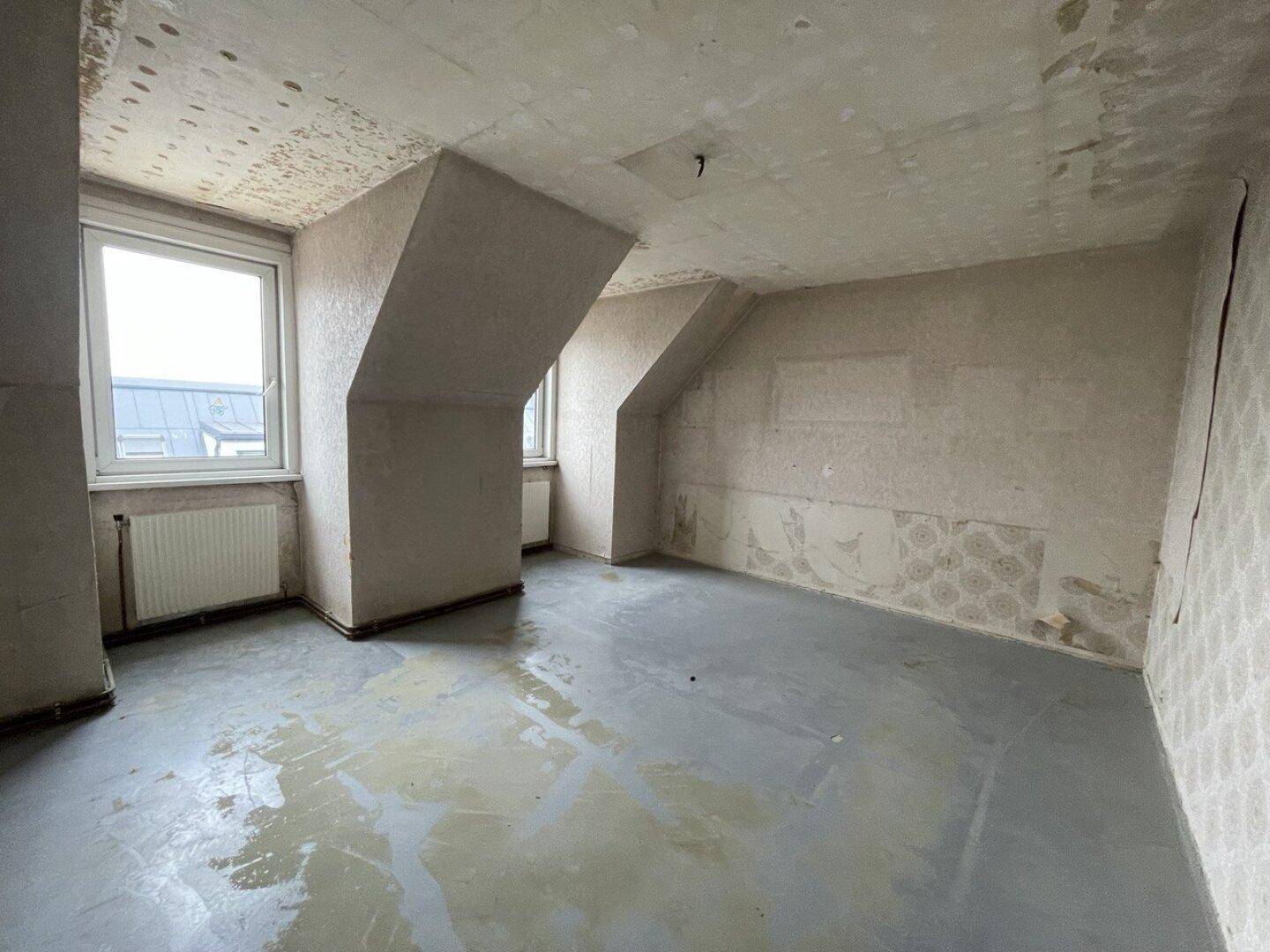 Unrenovierter Raum im Dachgeschoss mit schrägen Wänden, Fenster und Heizkörper, ideal für eine Neugestaltung.
