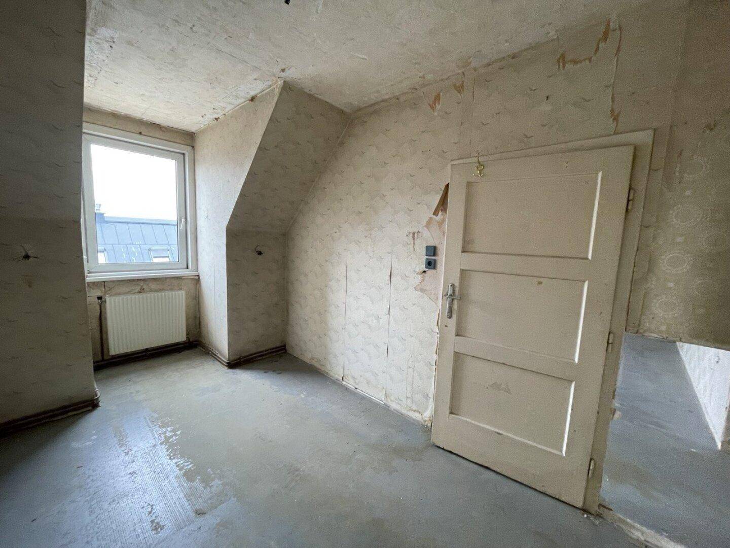 Unrenovierter Raum im Dachgeschoss mit Fenster, Heizkörper und abgenutzter Tapete, wartet auf Modernisierung.
