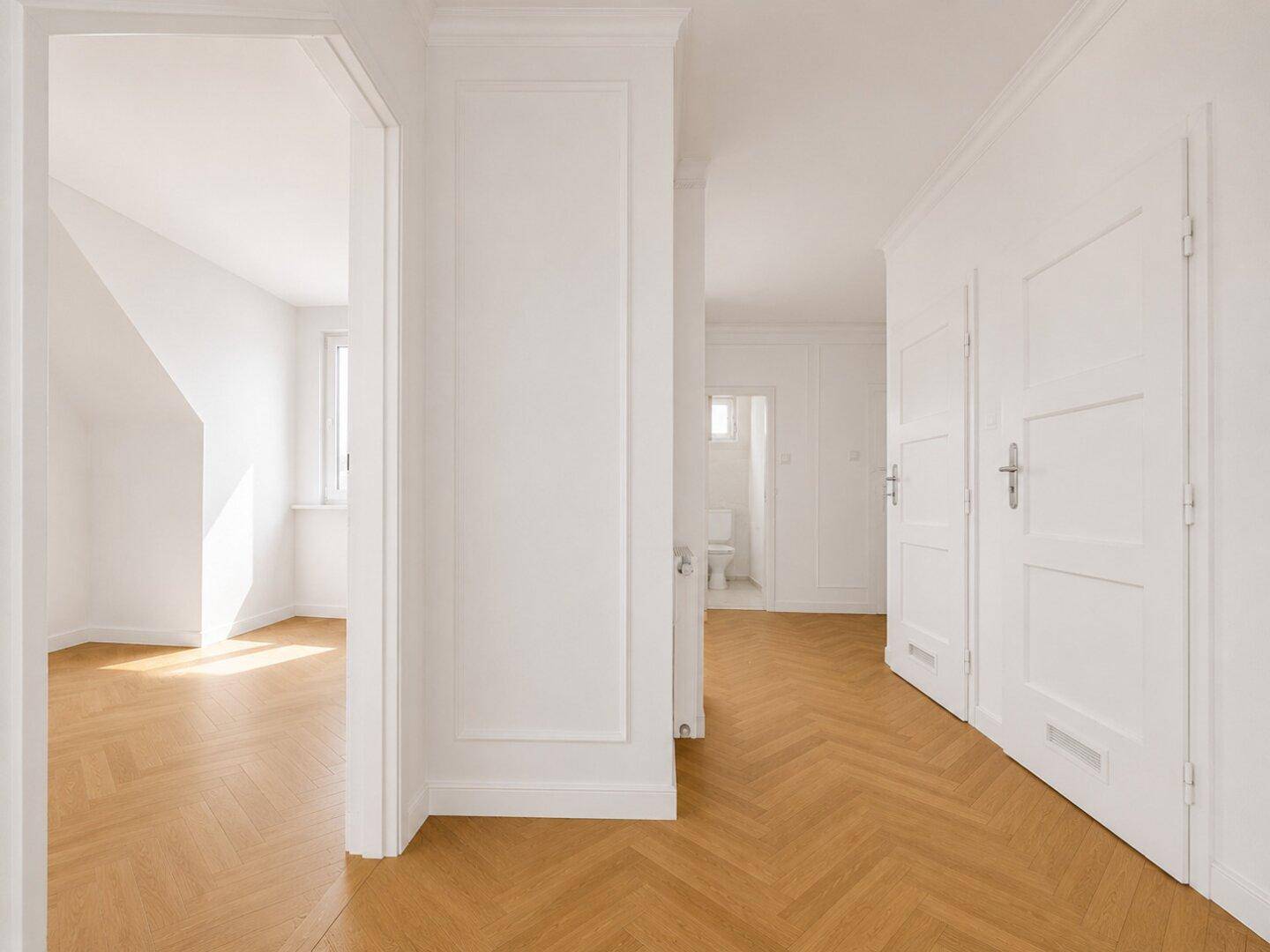 Heller, renovierter Flur mit Fischgrätparkett und weißen Wänden, führt zu weiteren Zimmern und einem WC.