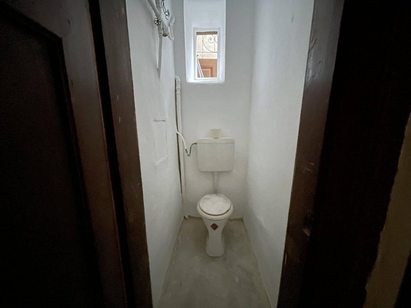 Kleine Toilette mit Fenster, weißer Keramik und einfacher Ausstattung in einem schmalen Raum.