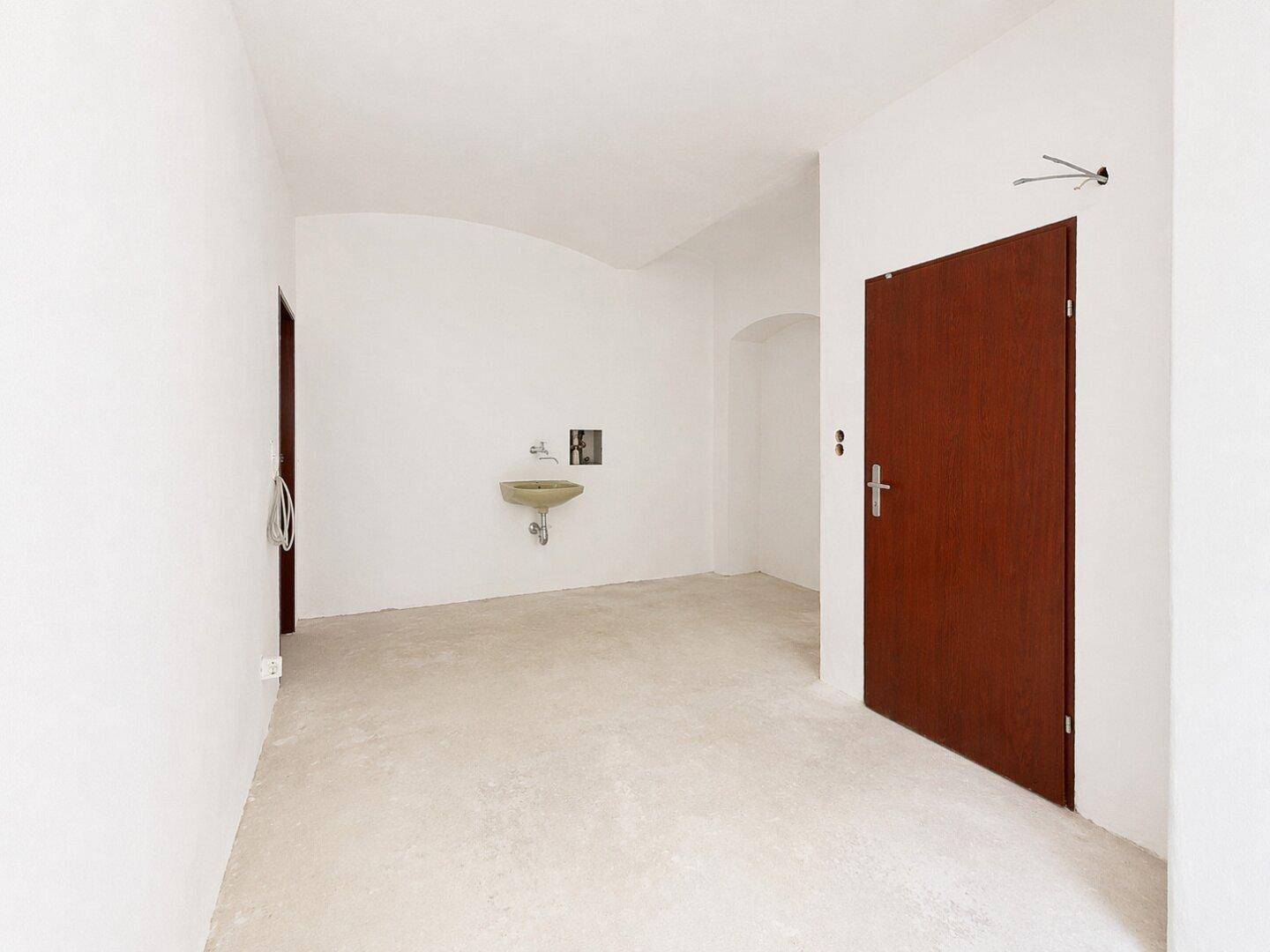 Heller, leerer Raum mit einem kleinen Waschbecken an der Wand und einer braunen Tür.