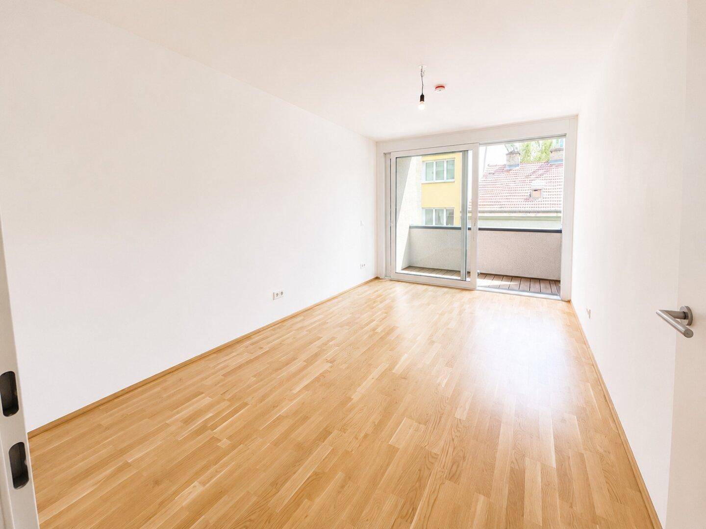 Helles, leeres Zimmer mit Holzboden und Schiebetür zum Balkon.