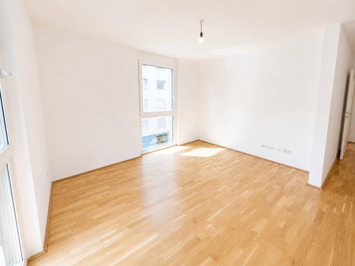 Helles, leeres Zimmer mit Holzboden und großem Fenster, ideal als Schlaf- oder Arbeitszimmer.
