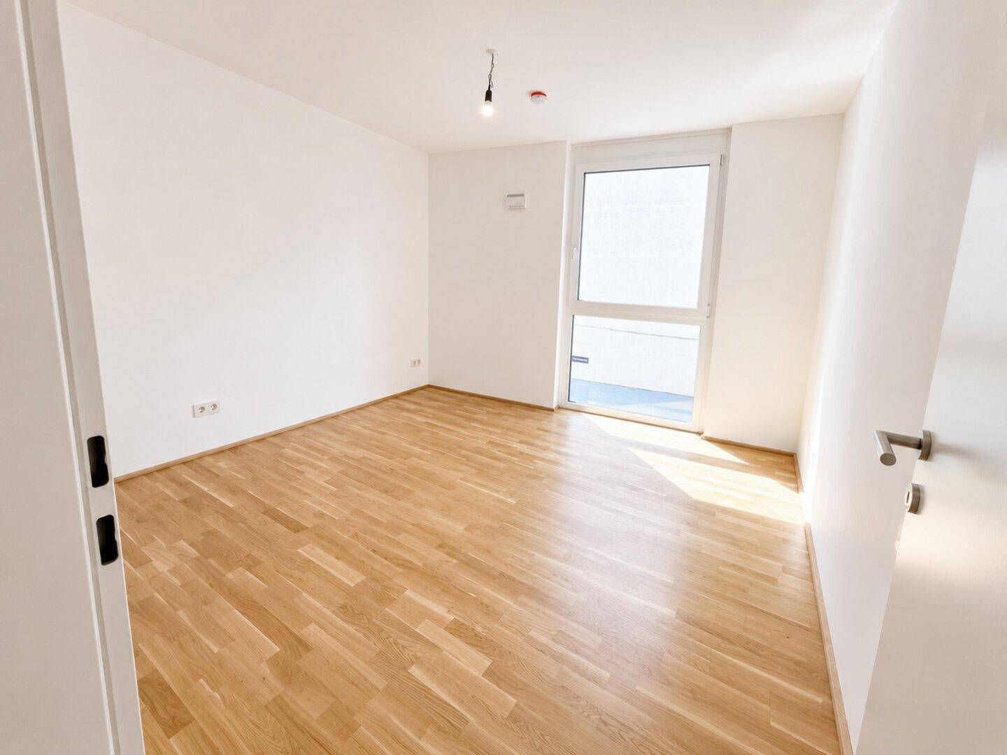 Helles, leeres Zimmer mit Holzboden und großem Fenster, bietet vielseitige Nutzungsmöglichkeiten.