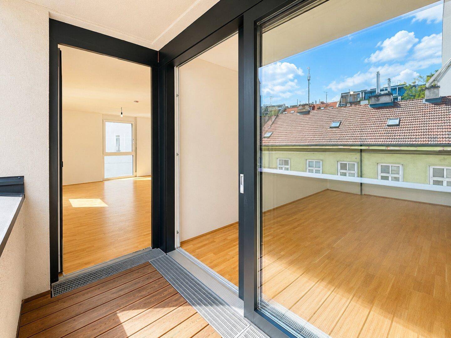 Blick vom Innenraum auf einen Balkon mit Holzboden und Dächern der Stadt.