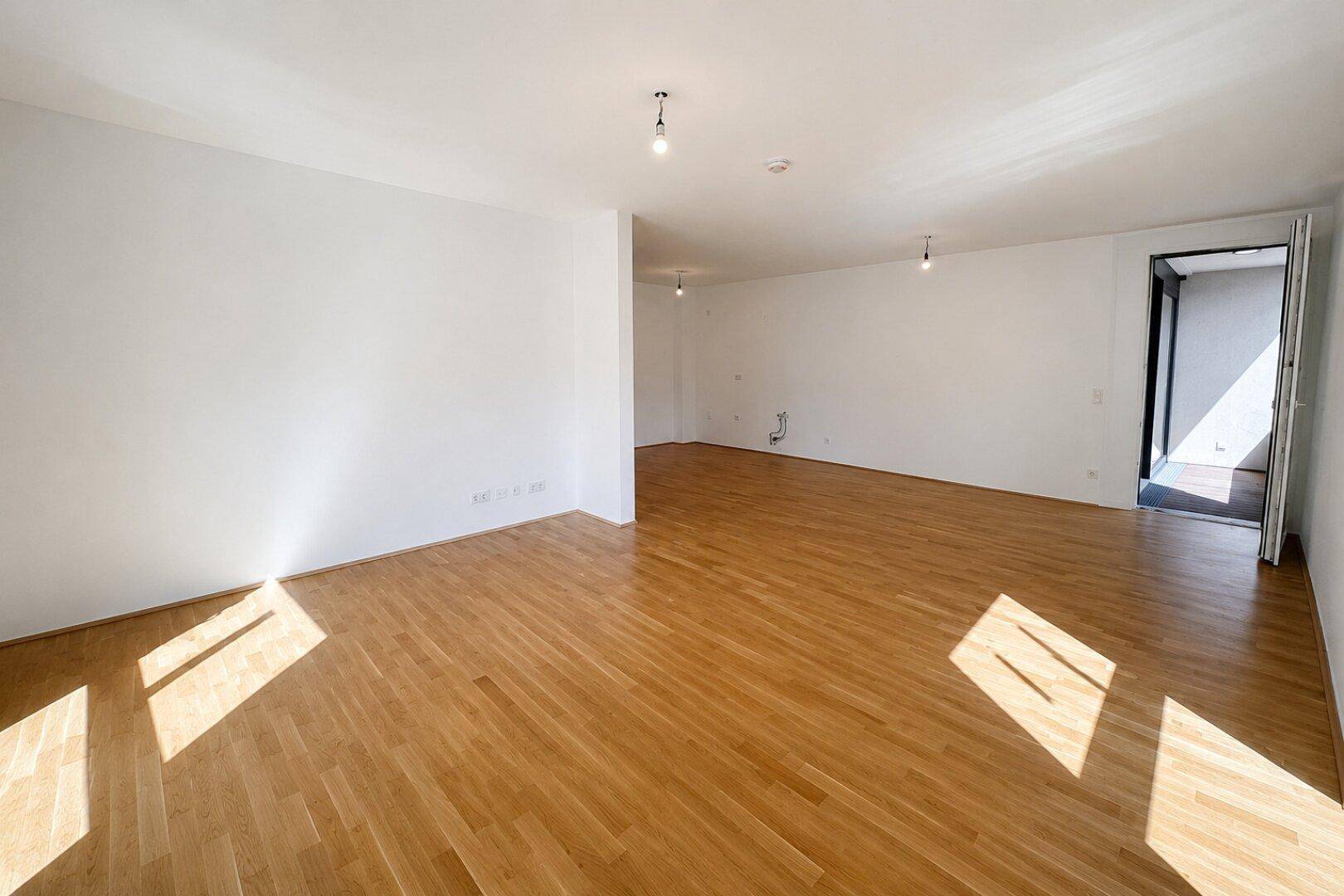 Geräumiger, leerer Wohnbereich mit Holzboden und Zugang zu einem Balkon.