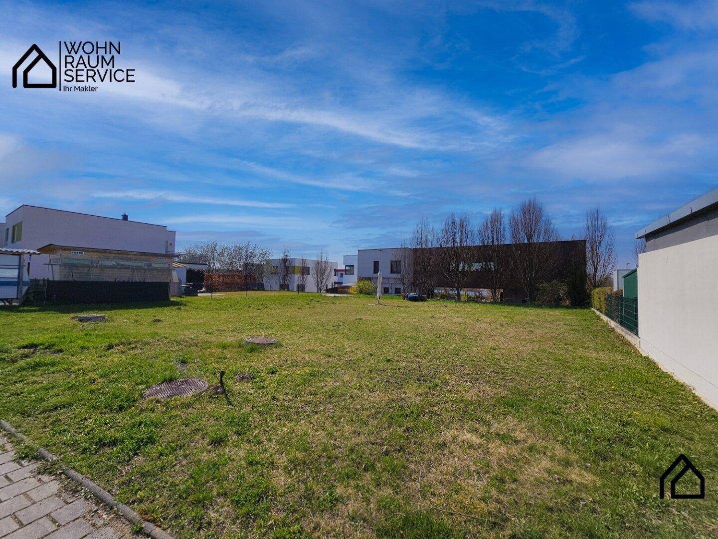 Eine freie, grüne Parzelle mit Gras und einigen Gebäuden im Hintergrund unter blauem Himmel.