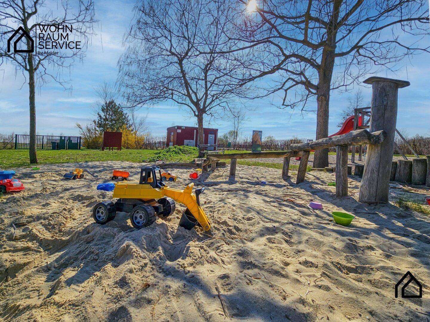 Ein großer Sandkasten mit Spielzeug und Holzspielgeräten unter strahlendem Sonnenschein.