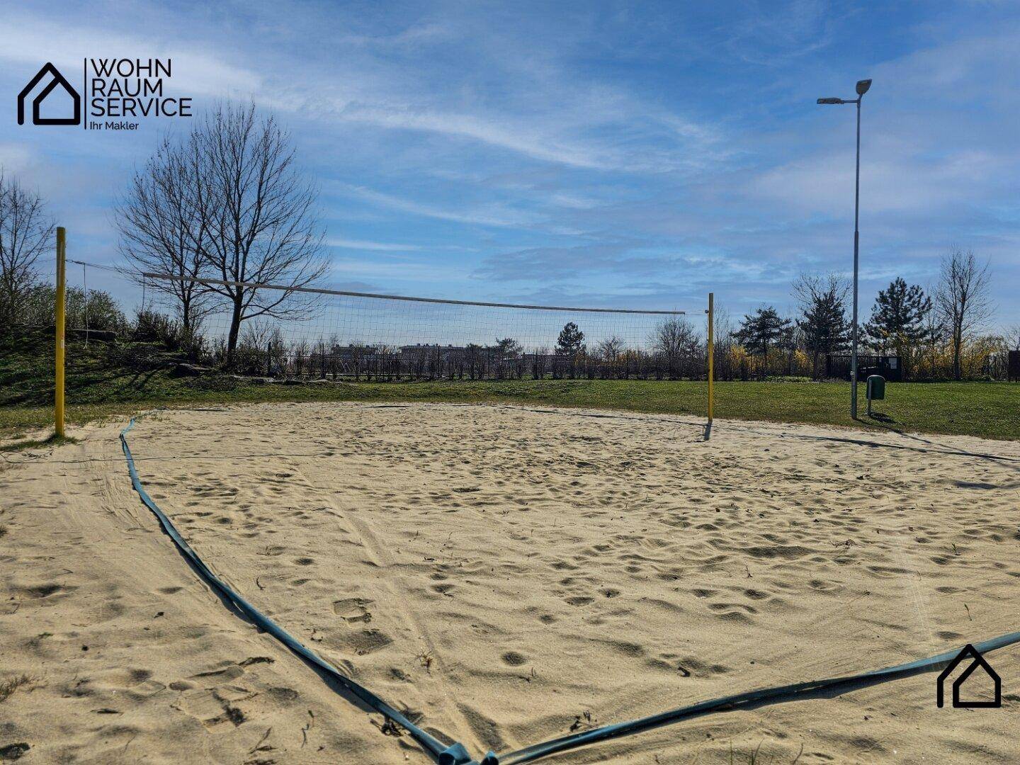 Ein Beachvolleyballfeld mit Netz und Sandboden, umgeben von Bäumen unter blauem Himmel.