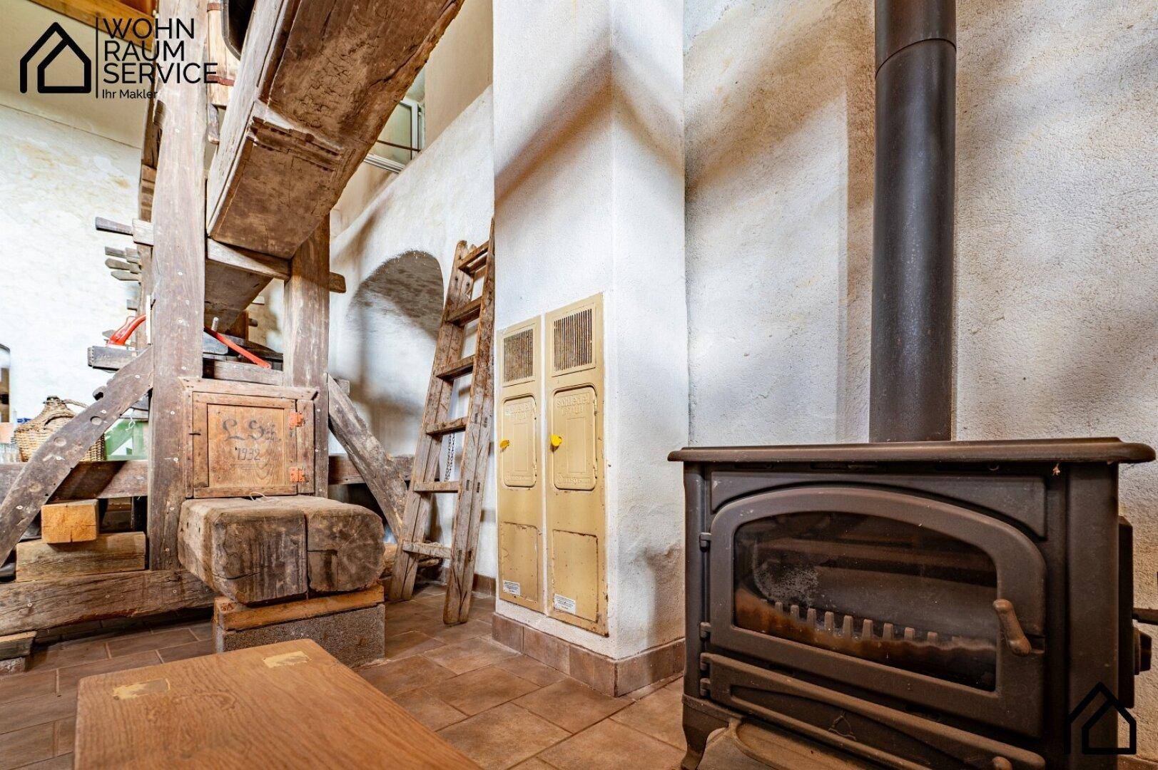 Alter Holzofen neben der historischen Weinpresse in einem rustikalen Raum mit Holzbalken.