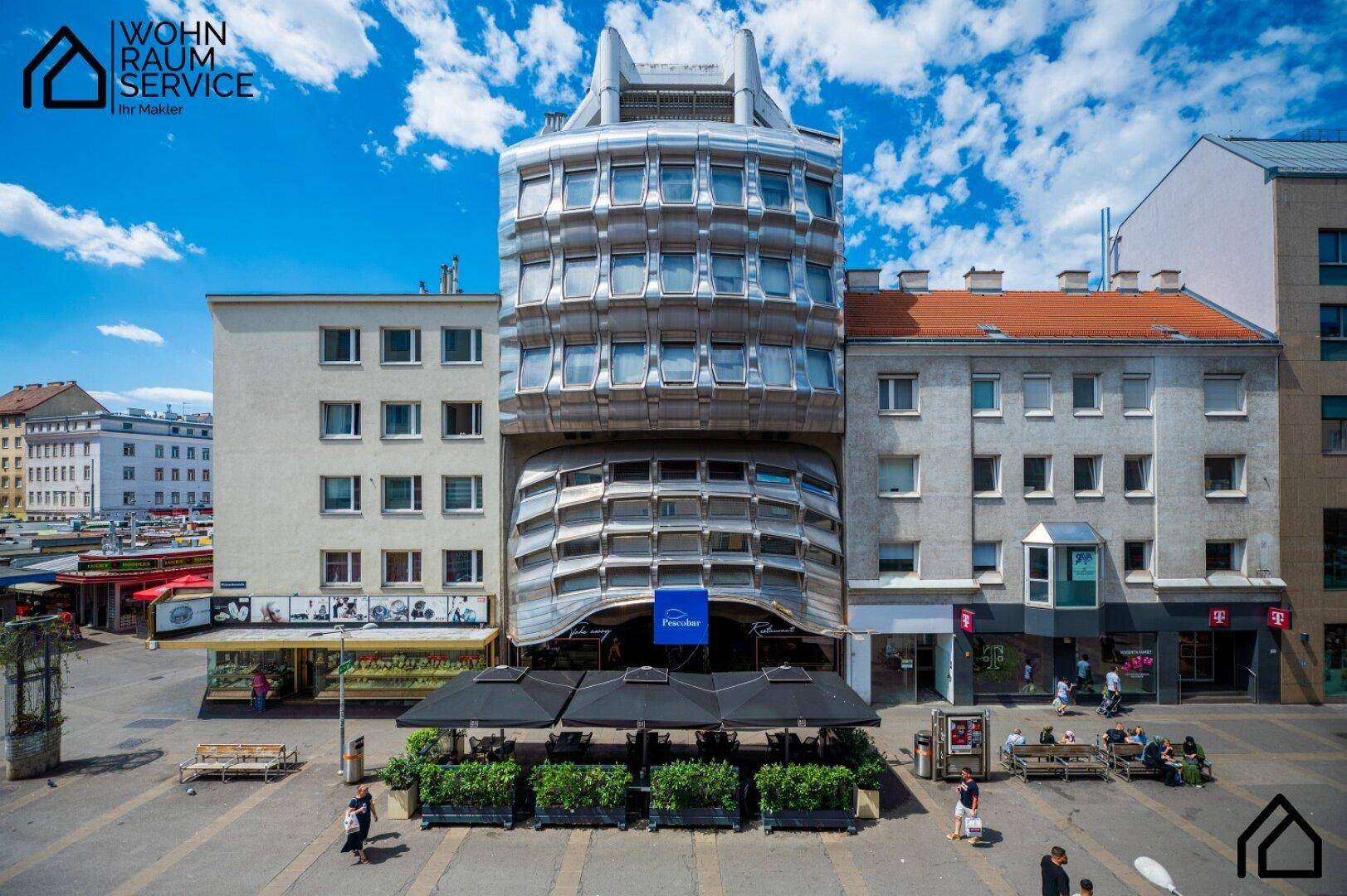 Auffällige Gebäudefassade an einem belebten Stadtplatz mit Cafés und Passanten.