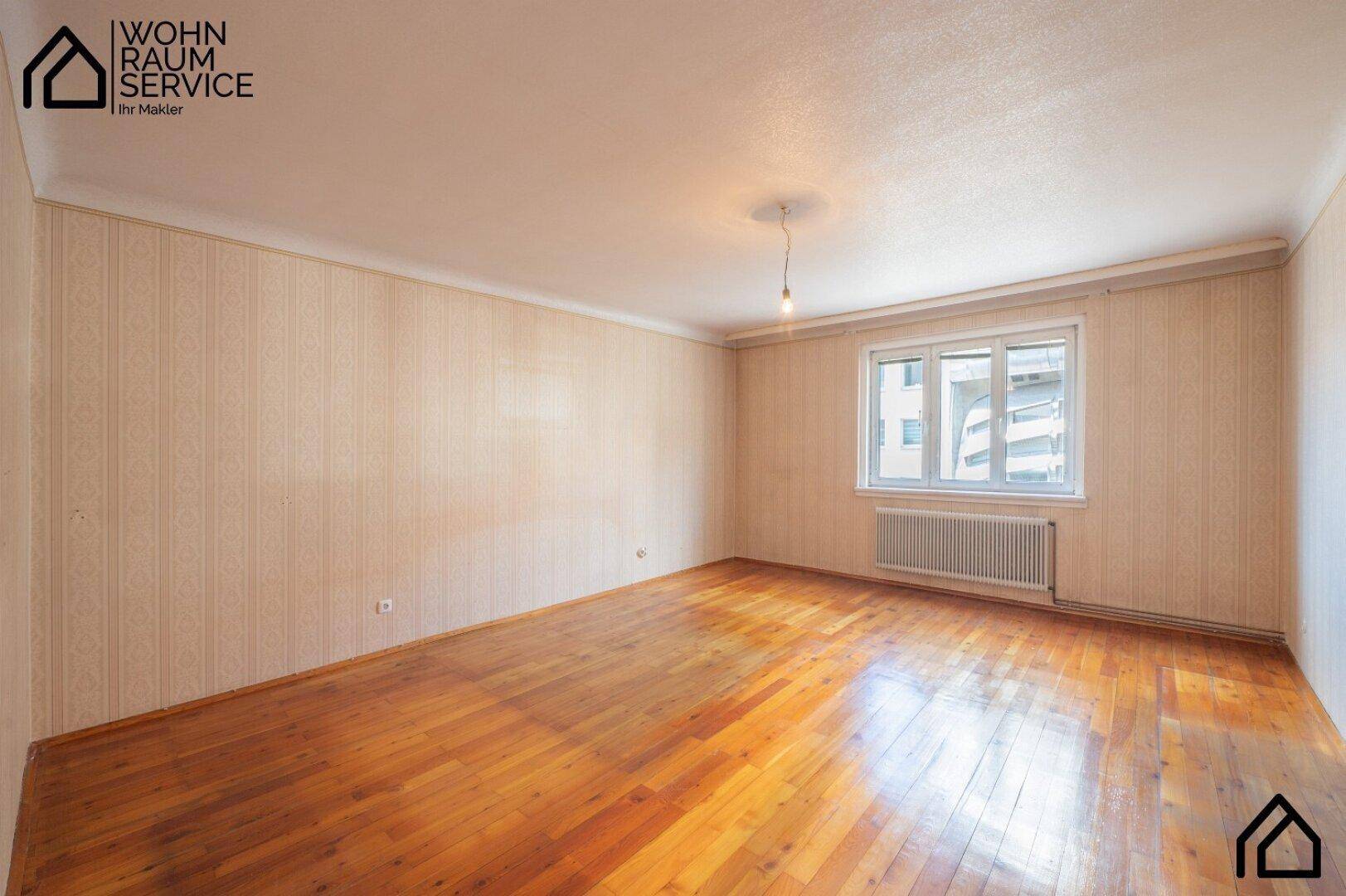 Leeres Zimmer mit hellem Holzparkett, gemusterter Tapete und Fenster.