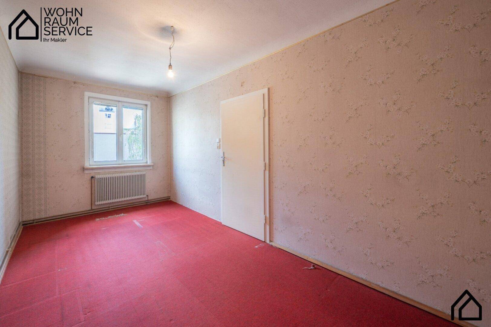 Leeres Zimmer mit rotem Teppichboden, gemusterter Tapete und Fenster.