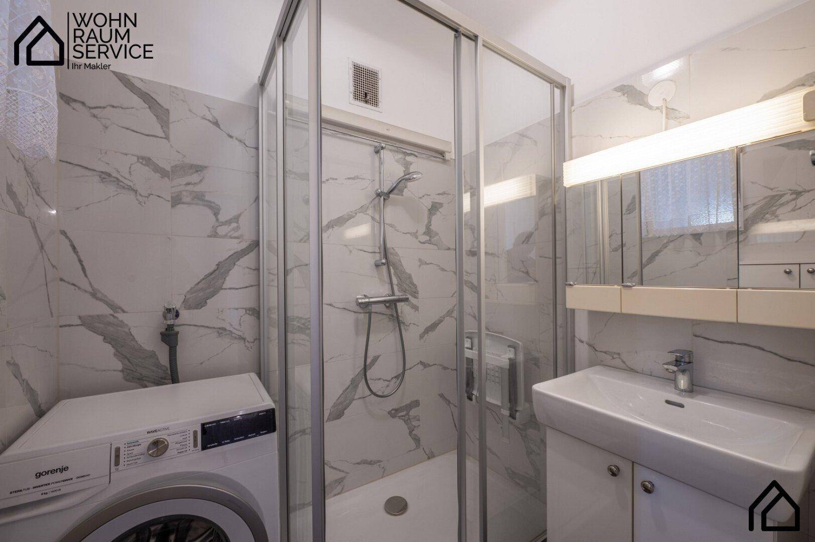 Modernes Badezimmer mit Duschkabine, Waschbecken und Waschmaschine, gefliest in Marmoroptik.