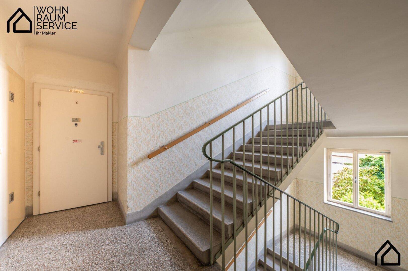 Helles Treppenhaus mit Fenster, Geländer und Terrazzoboden in einem Altbau.