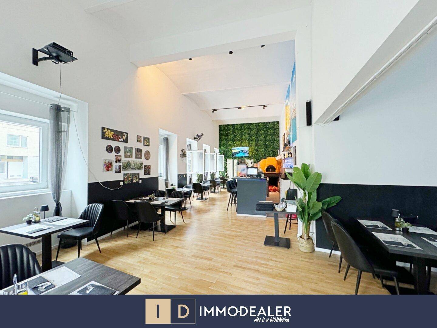 Weitläufiger Restaurantraum mit verschiedenen Sitzbereichen, hohen Decken und einem Pizzaofen im Hintergrund.