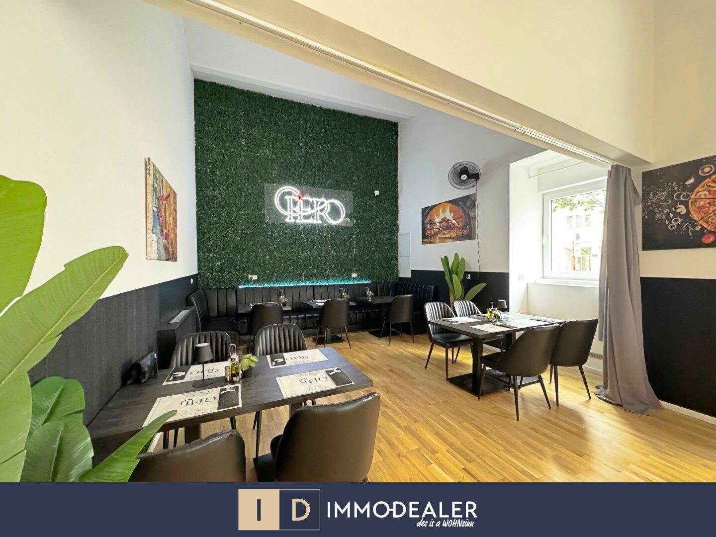 Heller Restaurantbereich mit Sitzbänken, Tischen und einer auffälligen grünen Pflanzenwand.