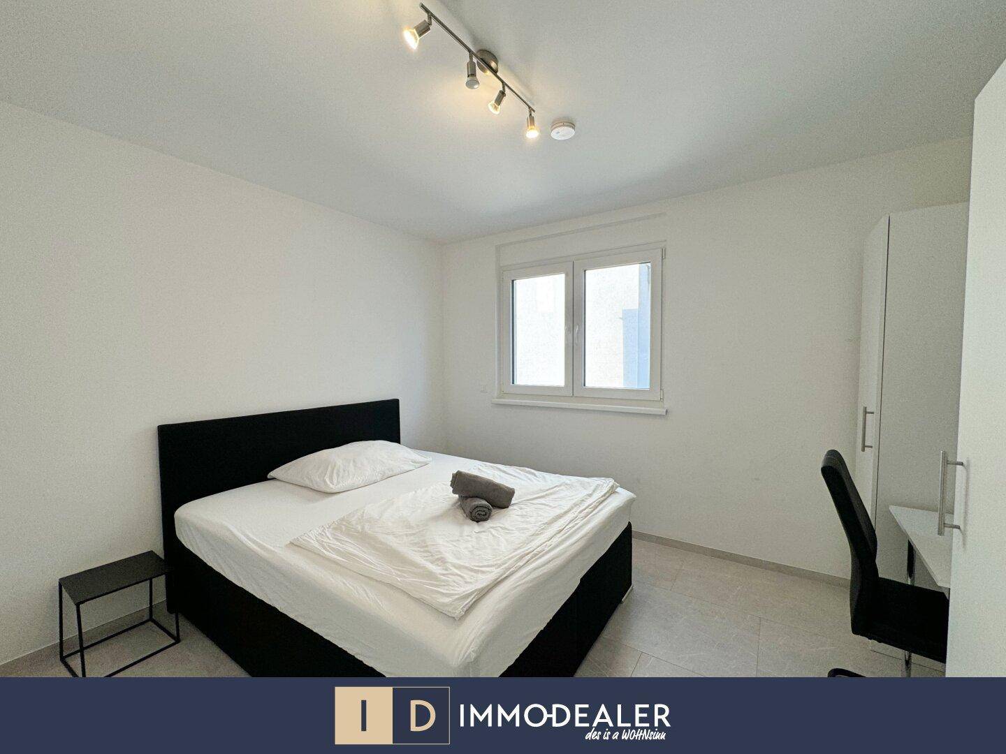 Modernes Schlafzimmer mit Doppelbett, Schreibtisch und Fenster, in heller Farbgebung gehalten.