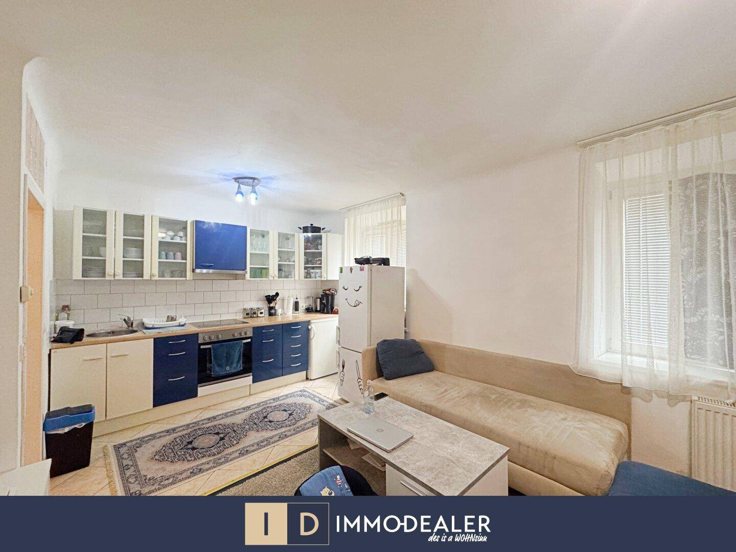 Heller Wohnbereich mit integrierter Küche, Sofa und Fenster mit Vorhängen.