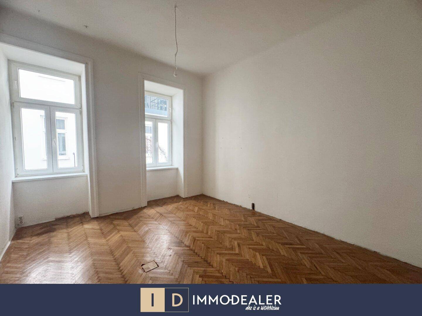 Geräumiges Zimmer mit Fischgrätparkett und großen Fenstern, bereit zur Renovierung.