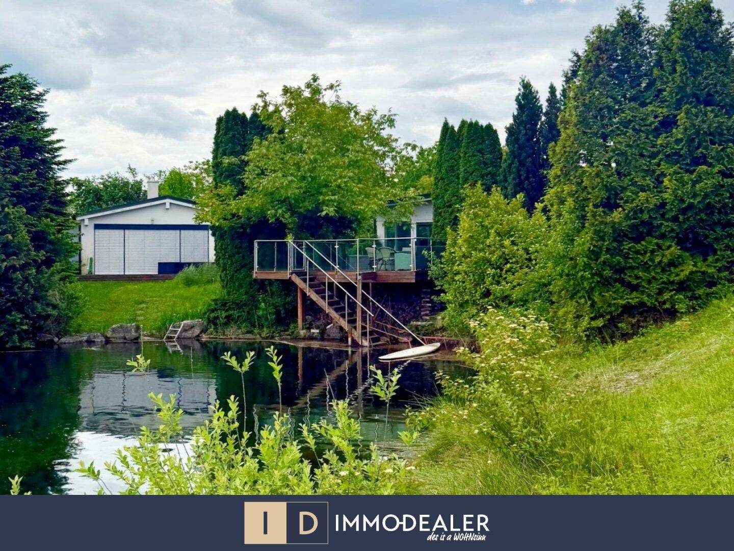 Haus am See mit großer Holzterrasse und Treppe zum Wasser, umgeben von viel Grün.