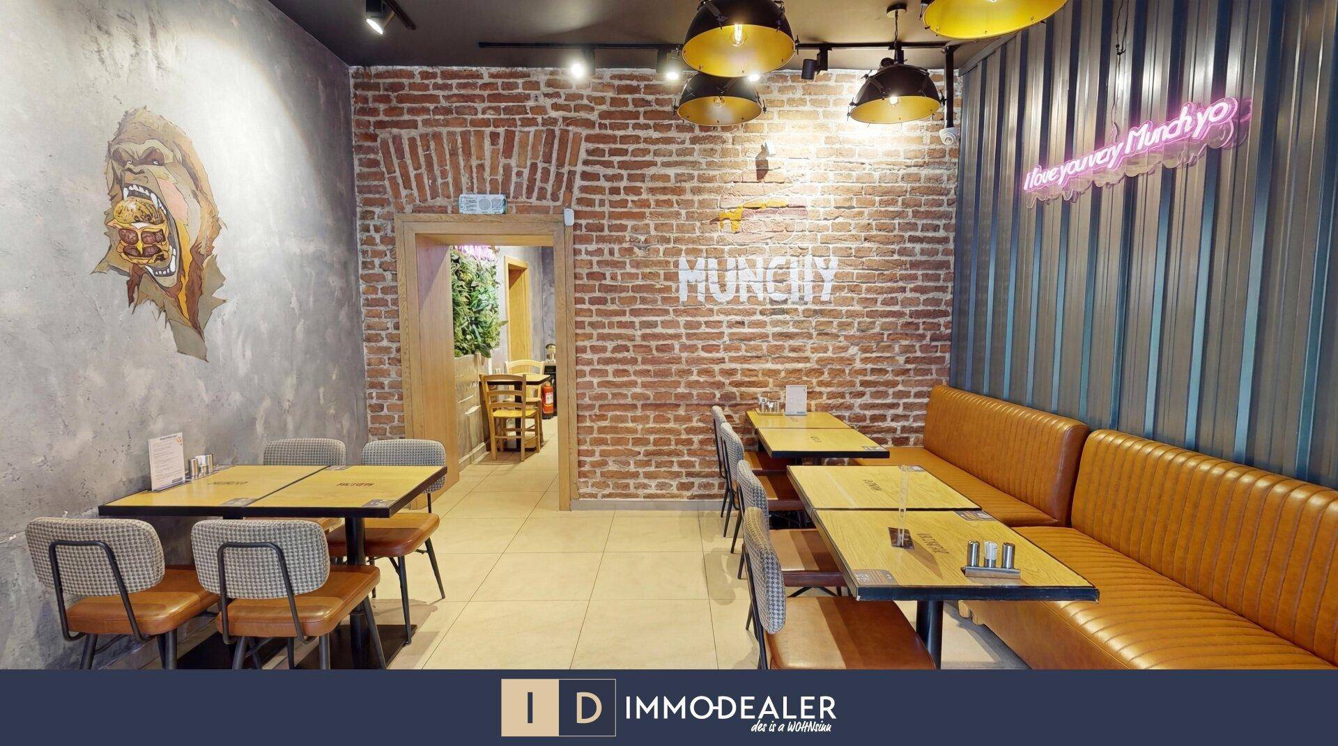 Geräumiger Restaurantbereich mit Backsteinwand, Sitzbänken und Tischen, beleuchtet von industriellen Hängelampen.