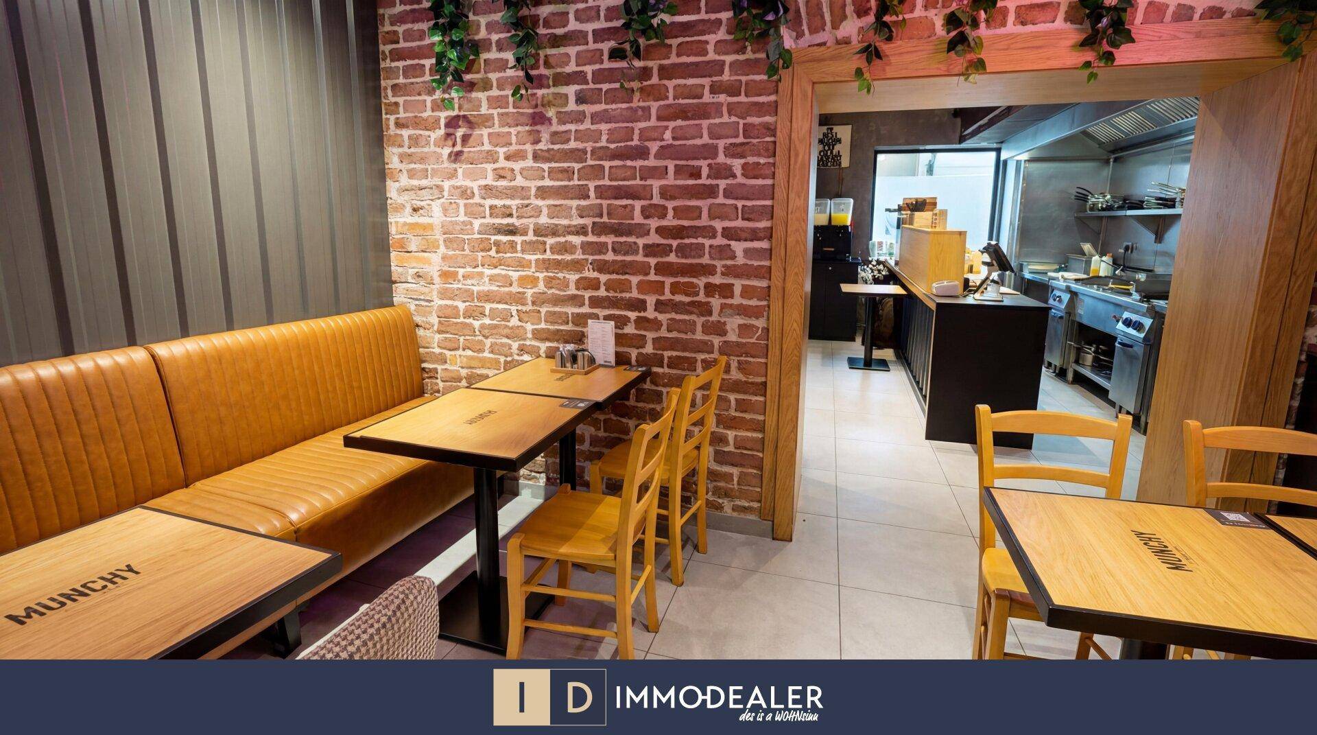 Restaurant-Sitzbereich mit Backsteinwand und Durchgang zur professionellen Küche, ausgestattet mit Holztischen.