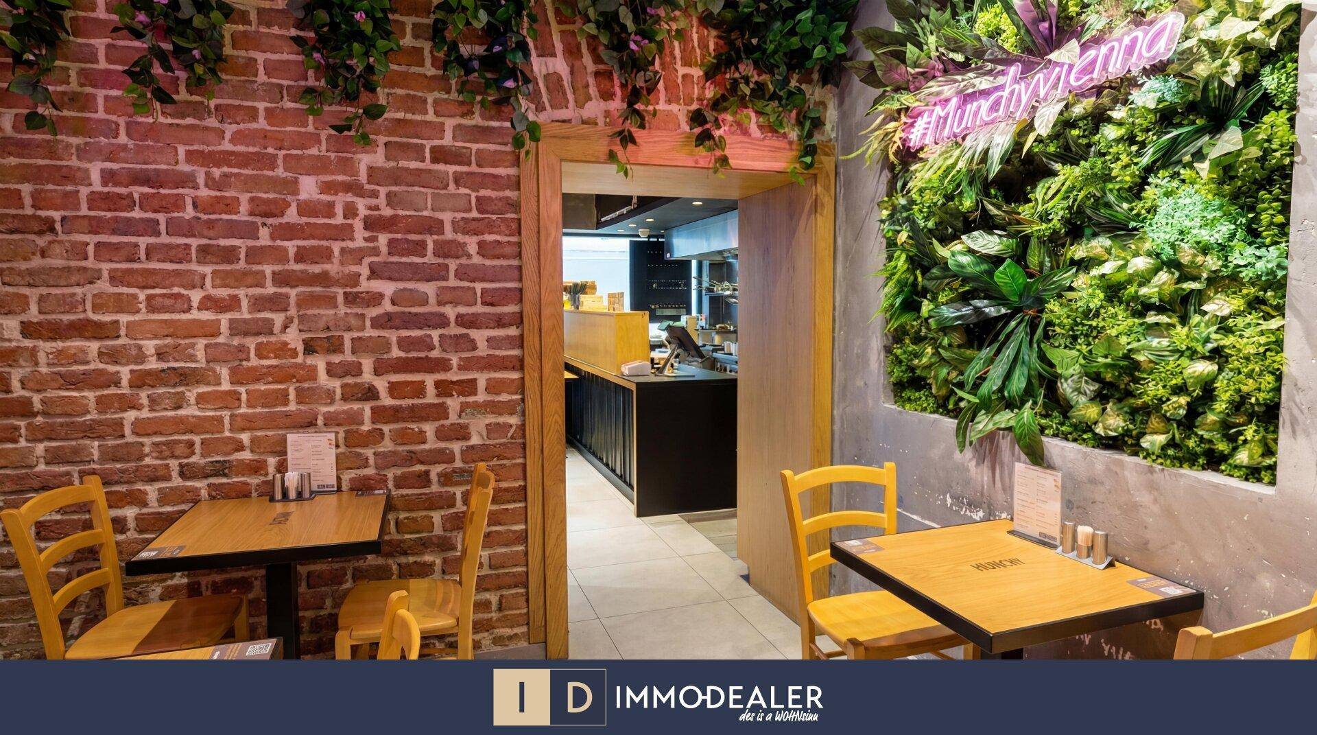 Restaurantbereich mit dekorativer grüner Wand, Backsteinmauer und Holztischen, die eine natürliche Atmosphäre schaffen.