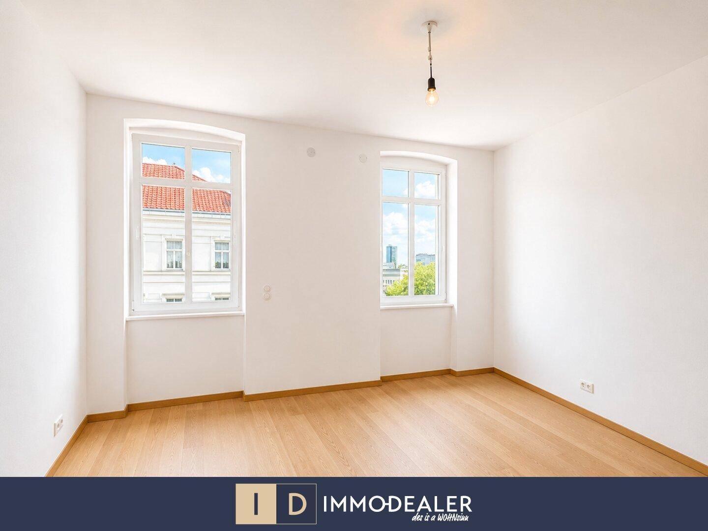 Helles Zimmer mit Parkettboden und zwei großen Fenstern, die viel Tageslicht hereinlassen.