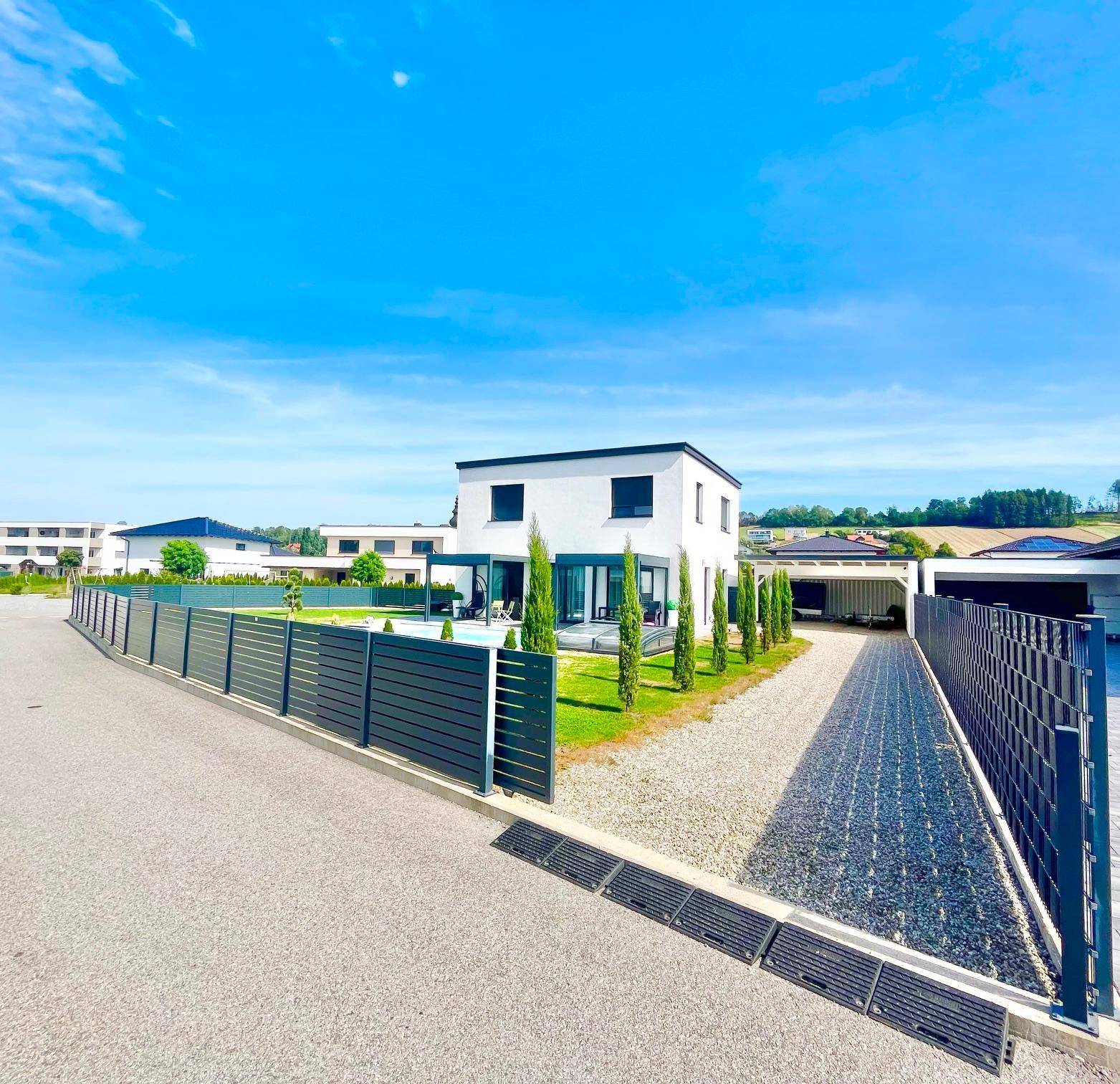 Weitläufige Seitenansicht des Hauses mit Kiesauffahrt, Zaun und dem dahinterliegenden Poolbereich.