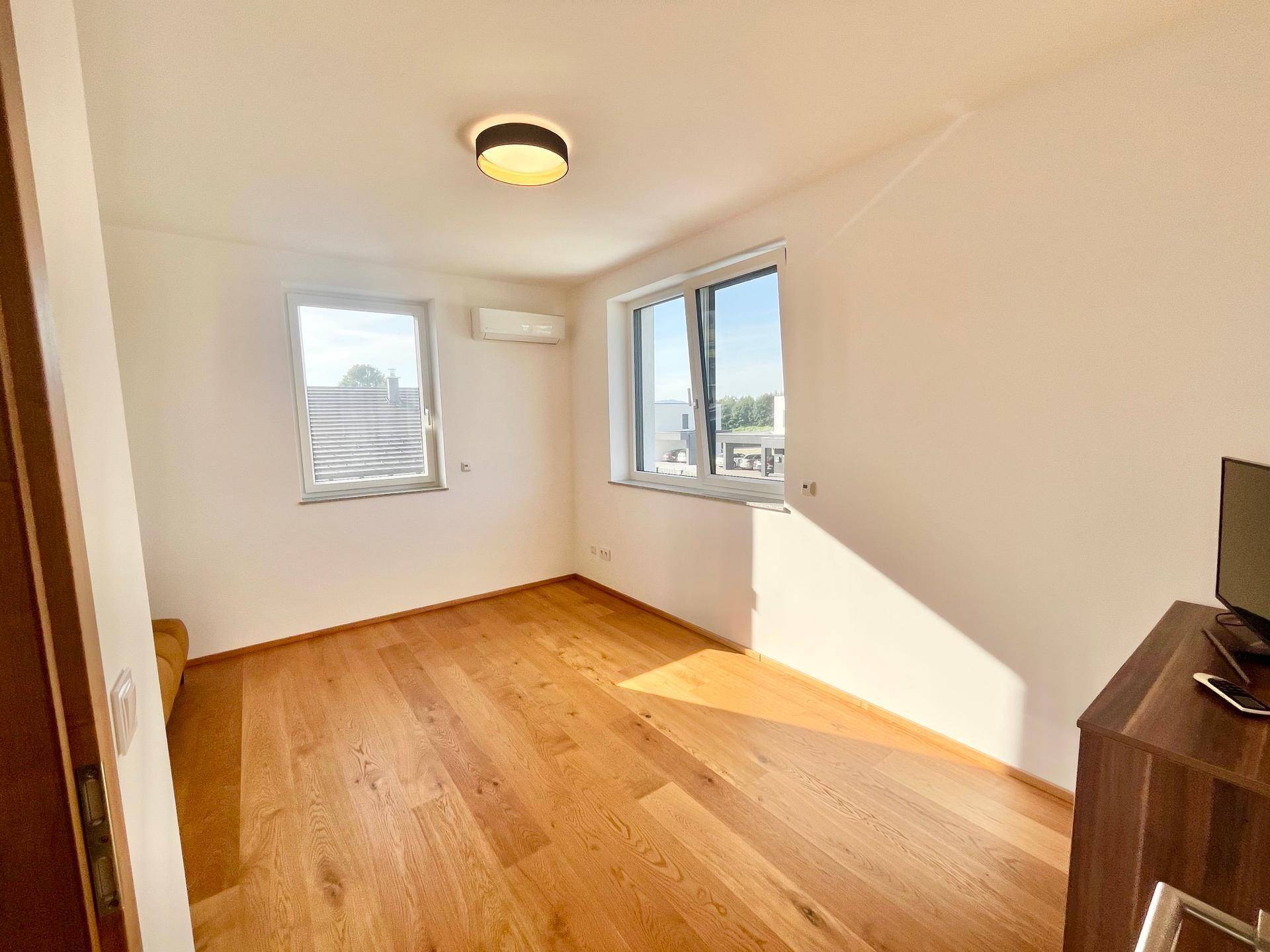 Sonniger Schlafbereich mit Holzboden, weißen Wänden und zwei Fenstern.
