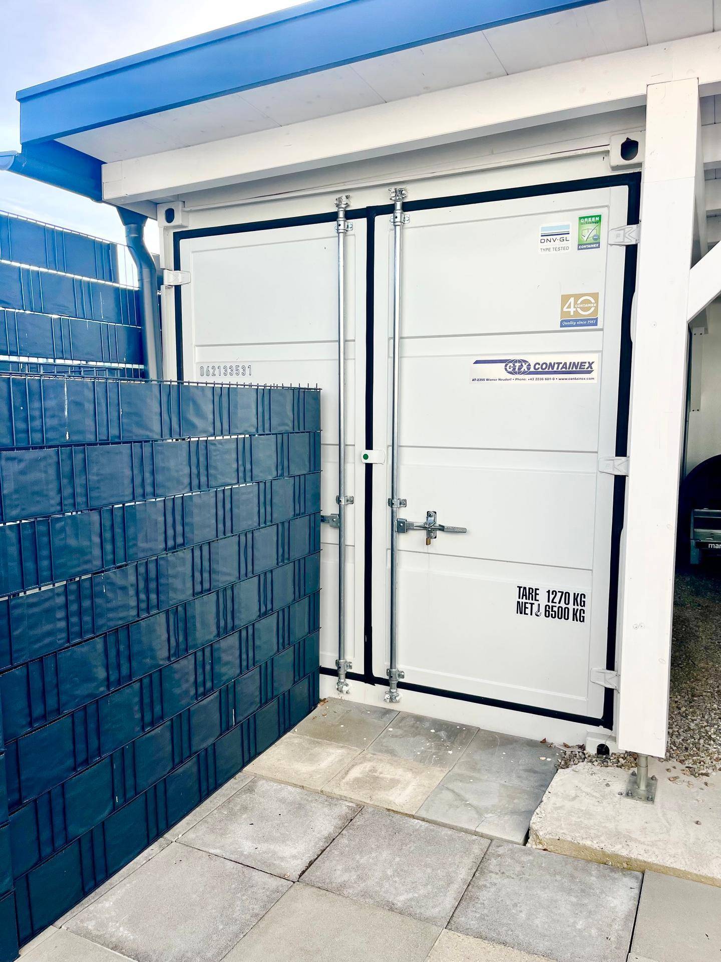 Weißer Lagercontainer neben einem modernen Zaun, dient als zusätzliche Abstellmöglichkeit.
