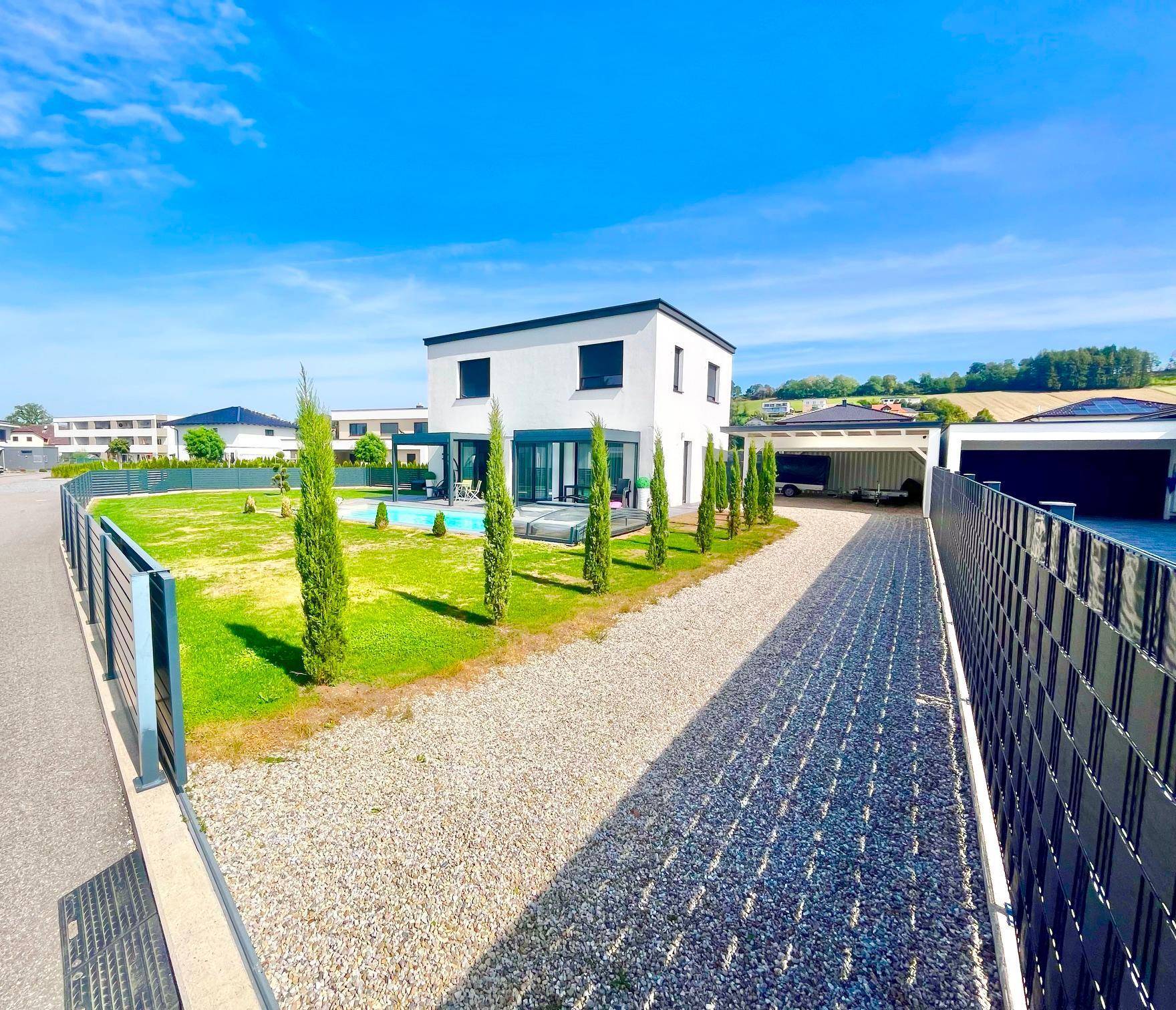 Seitenansicht des modernen Hauses mit Kiesauffahrt, Zypressenreihe und Blick auf den Poolbereich.