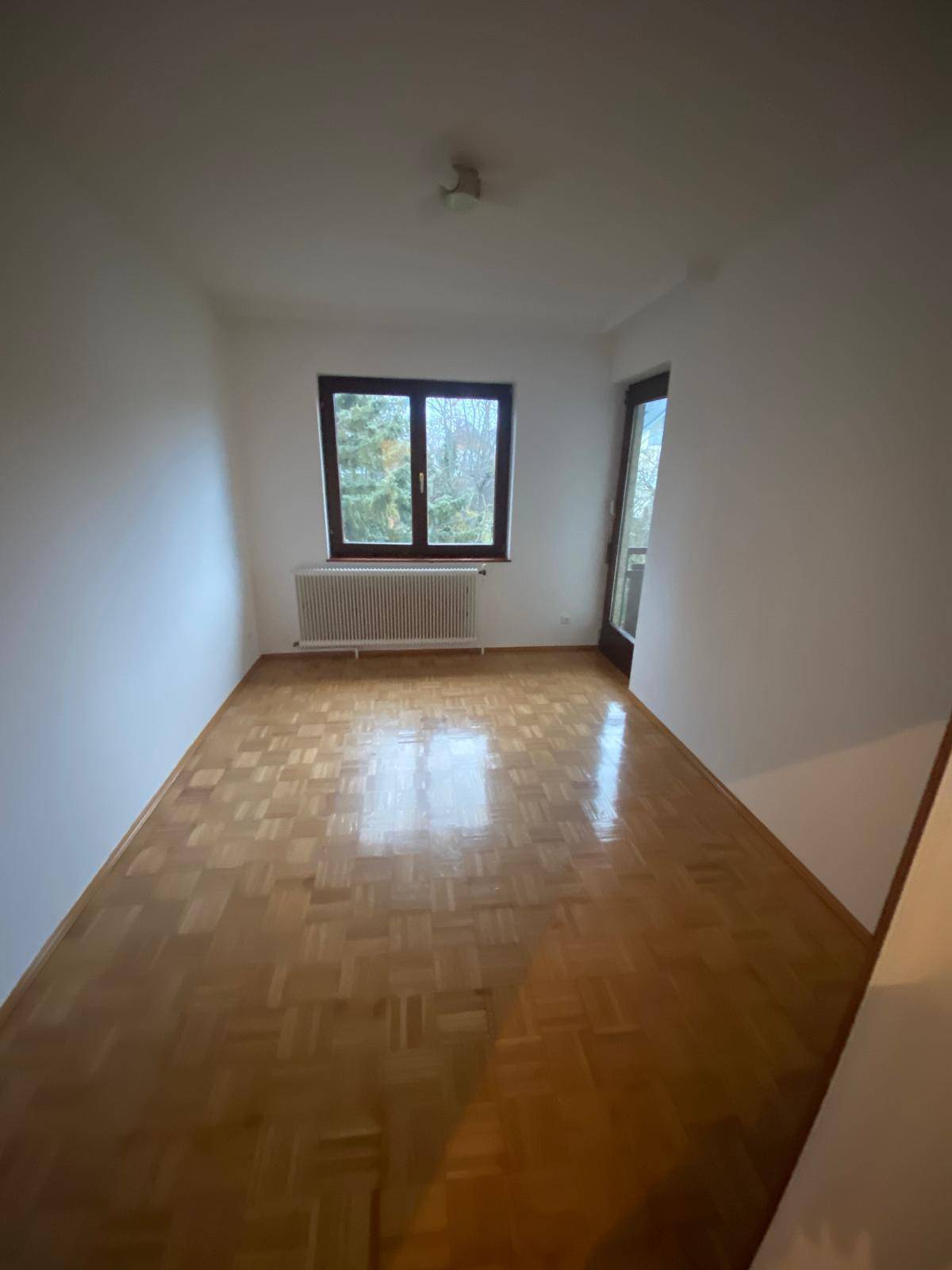Helles, leeres Zimmer mit Parkettboden, Fenster und Heizkörper.