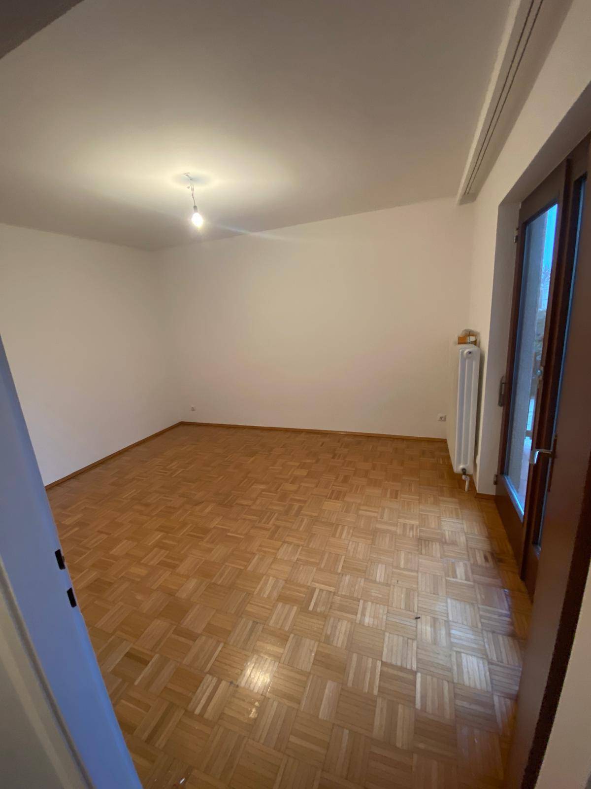 Geräumiges, leeres Zimmer mit Parkettboden und großem Fenster, das viel Licht hereinlässt.