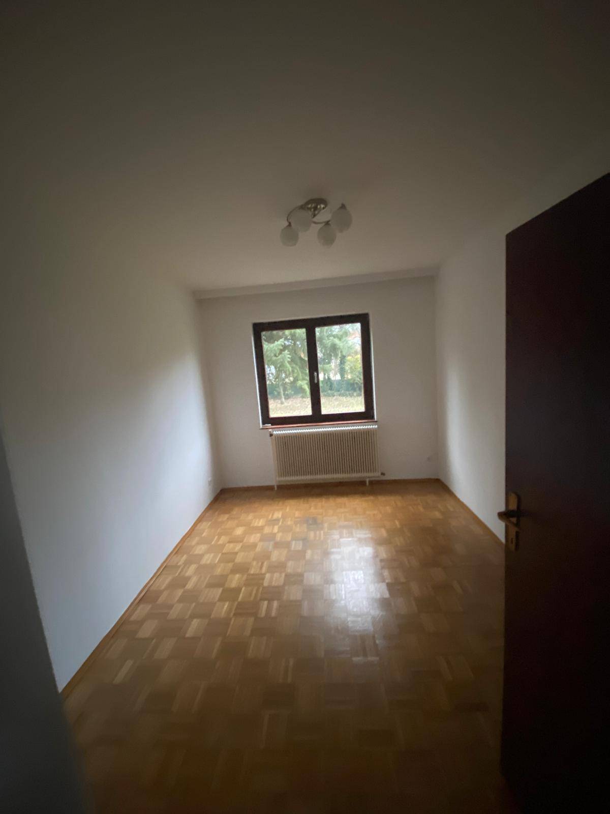 Helles, leeres Zimmer mit Parkettboden, Fenster und Heizkörper.