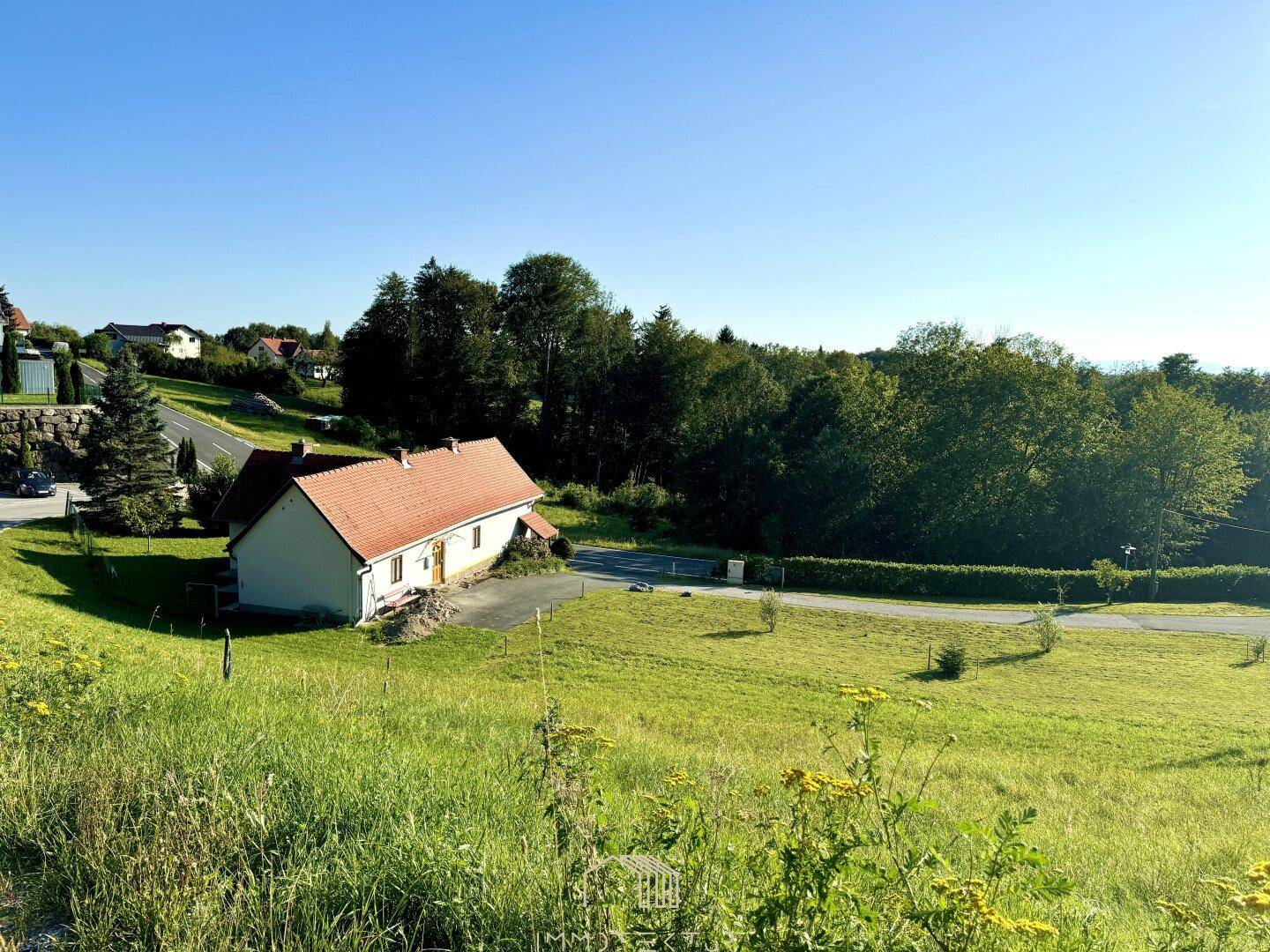 Ländliche Szenerie mit einem traditionellen Haus, rotem Dach und weitläufigen grünen Wiesen unter blauem Himmel.