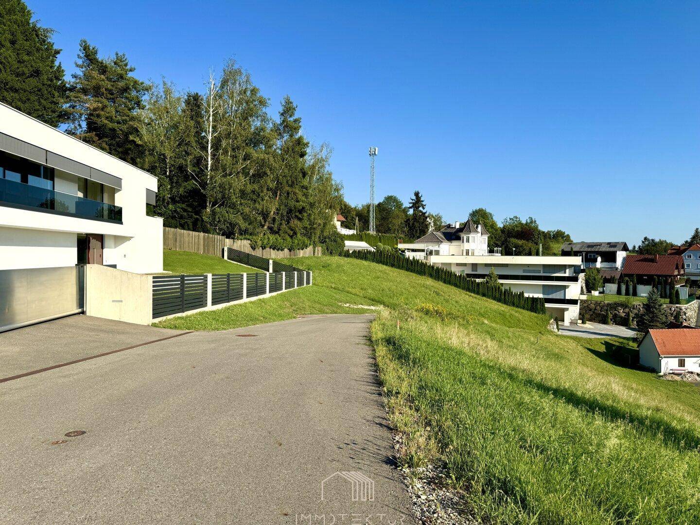Moderne Architektur mit weißer Fassade, Garagenzufahrt und einem Zaun an einem grünen Hang.