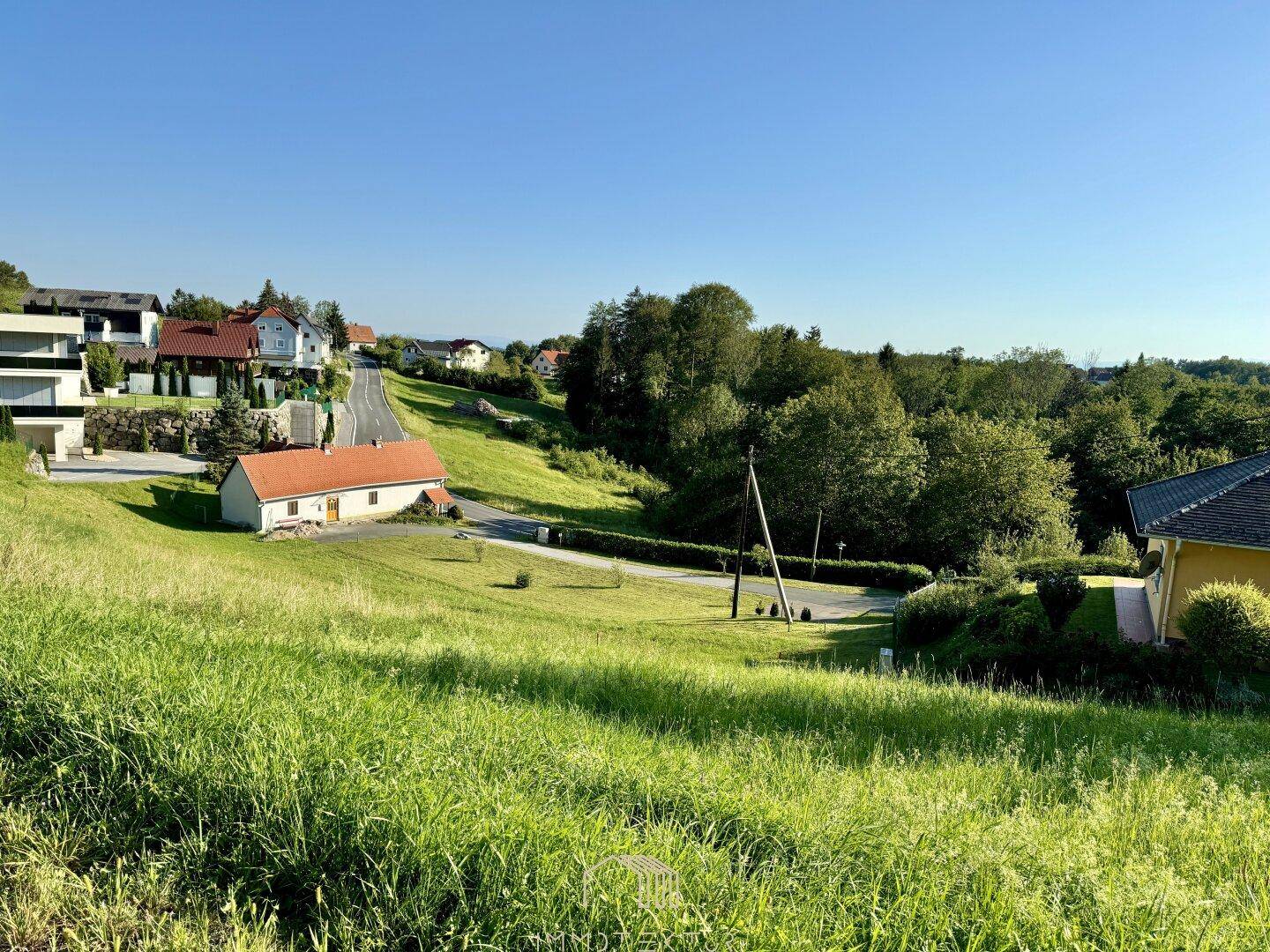 Weitläufige Landschaft mit grünen Hügeln, Wohnhäusern und einer Straße unter klarem Himmel.