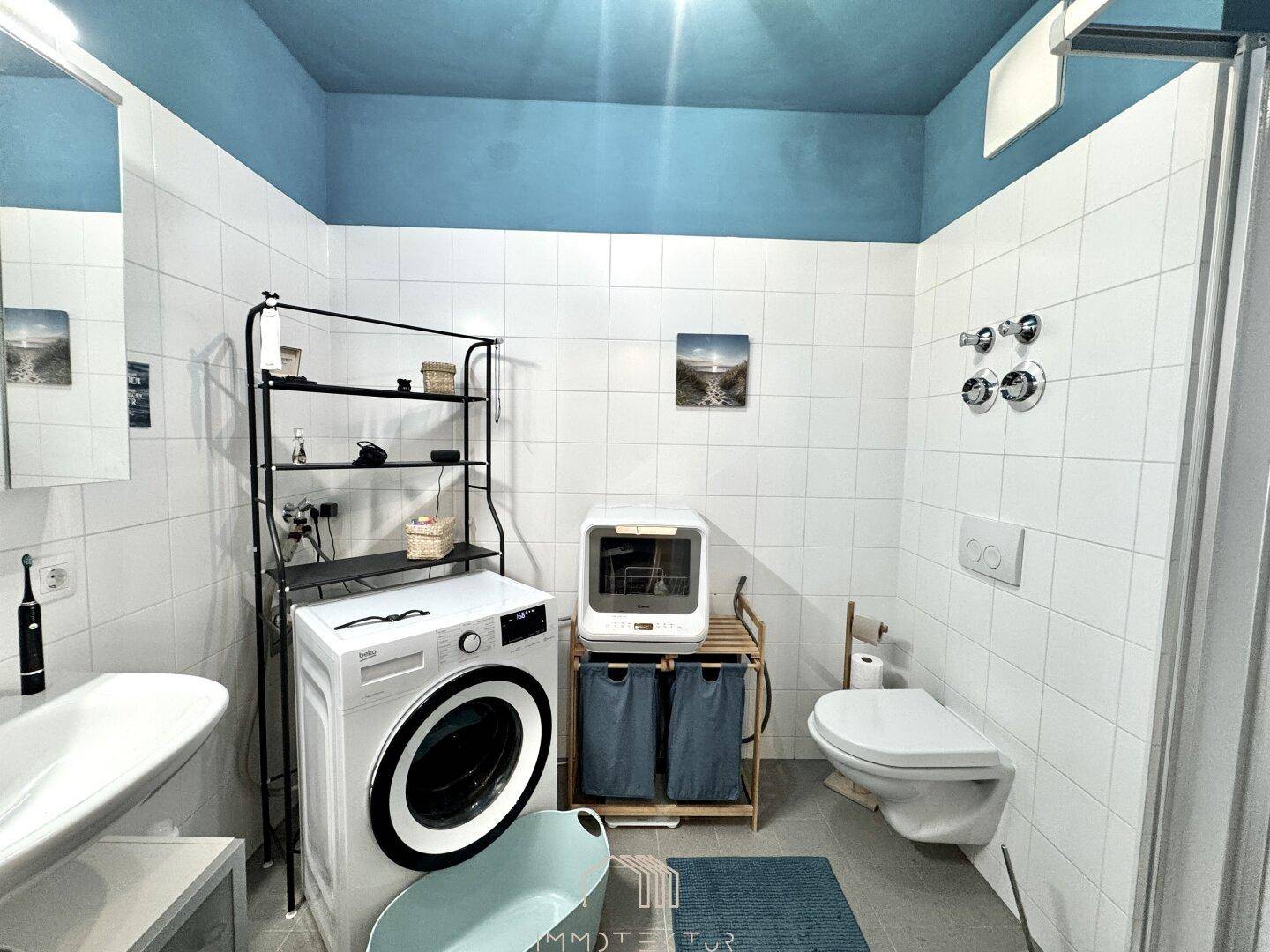 Funktionales Badezimmer mit Dusche, WC, Waschmaschine und weißen Fliesen.
