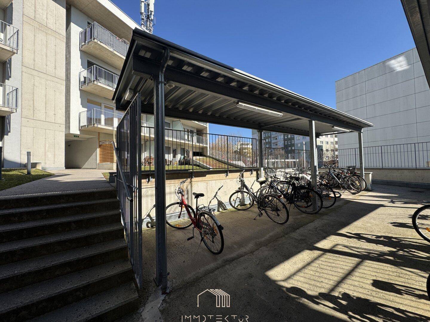 Überdachter Fahrradabstellplatz vor einem Mehrfamilienhaus an einem sonnigen Tag.