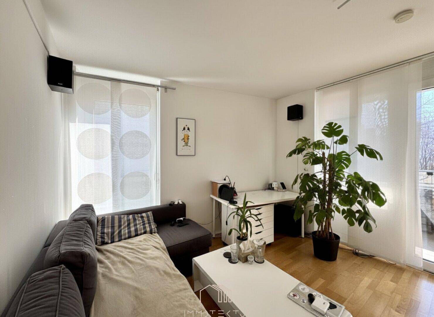 Heller Wohnbereich mit Sofa, Schreibtisch und Zugang zum Balkon.
